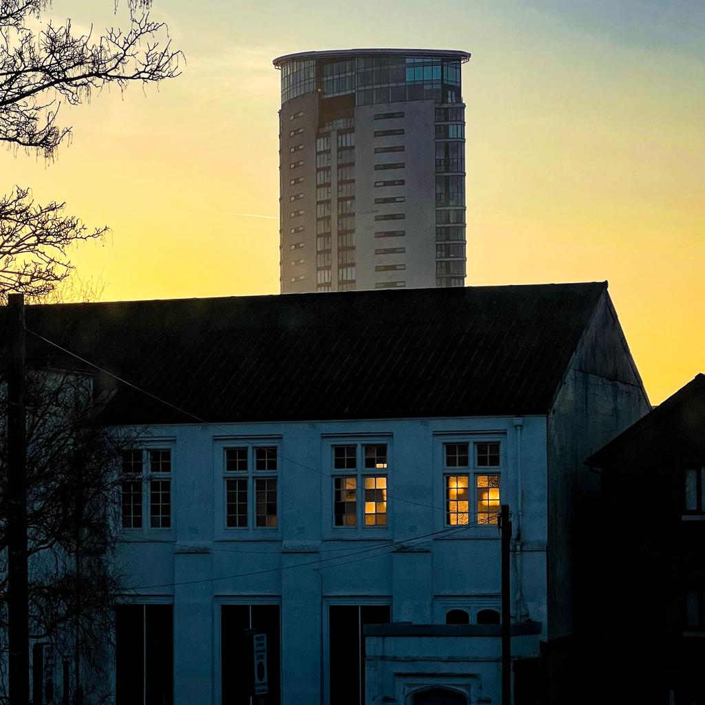 The sun rising beyond The Tower but still behind a building in the foreground. The sun shines through the windows of the upper floor of the foreground building.