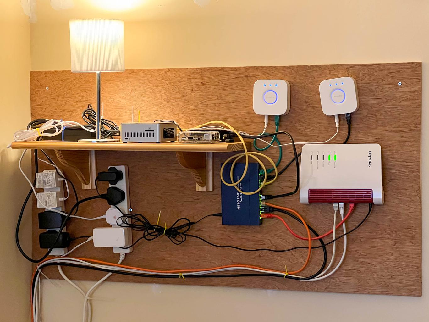 Wall-mounted networking electronics: router, network switch, a couple of Hue hubs; a Linux NUC, a couple of Pis, and a lamp on a shelf; two bars of electrical outlets and cables, lots of cables, many with zipties.