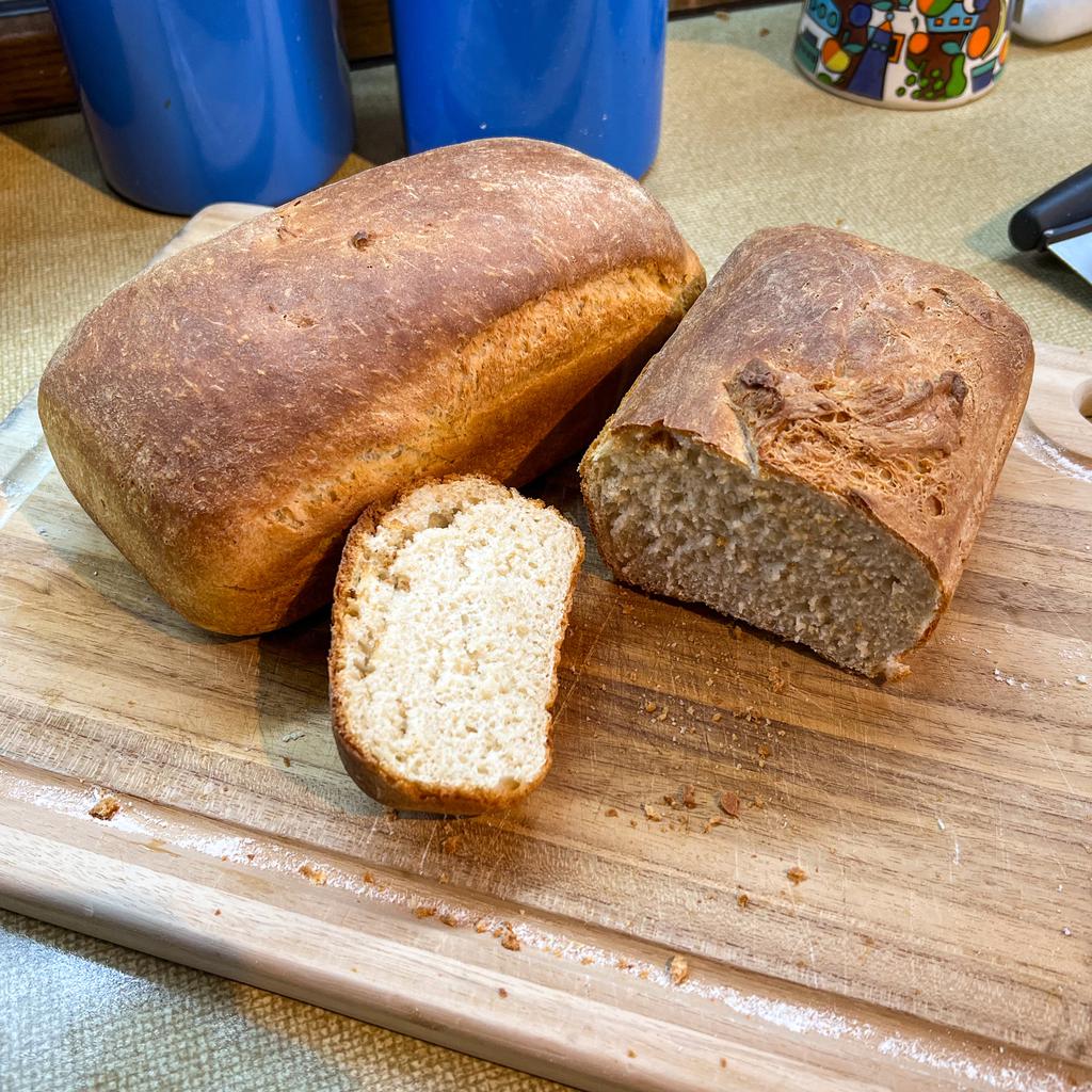 Two loaves of crusty white bread on a breadboard. One loaf cut open to reveal the inside.