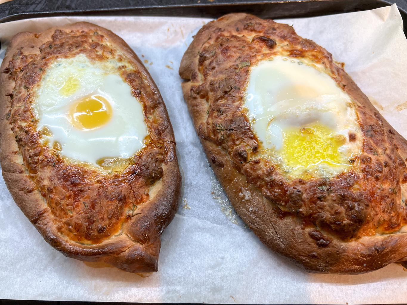 Two just baked khachapuri, savory pastry “boats” filled with cheese and herbs. Part way through cooking, a raw egg is added to the top and then baked until firm.