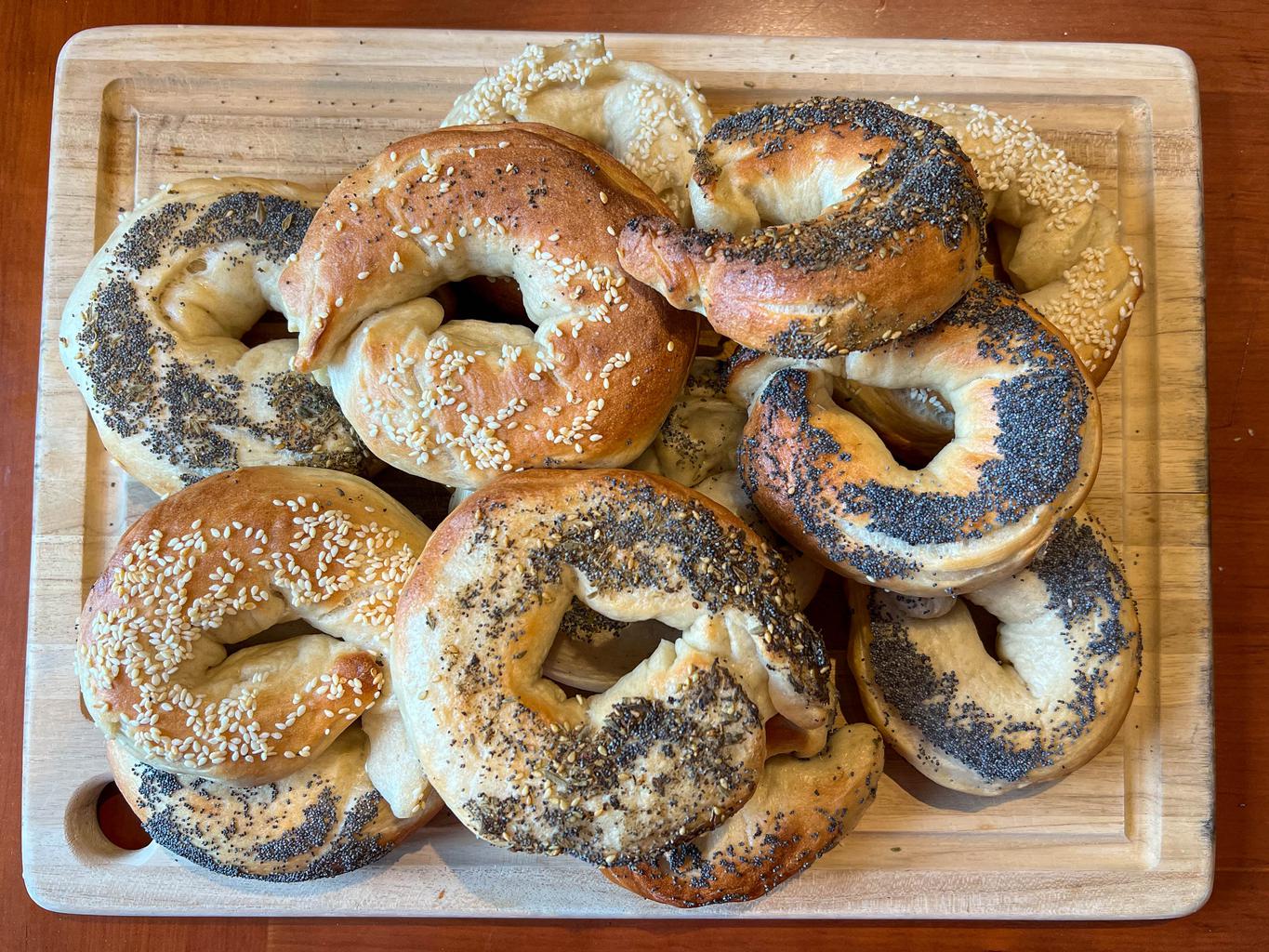 A dozen golden brown, baked bagels piled up on a bread board