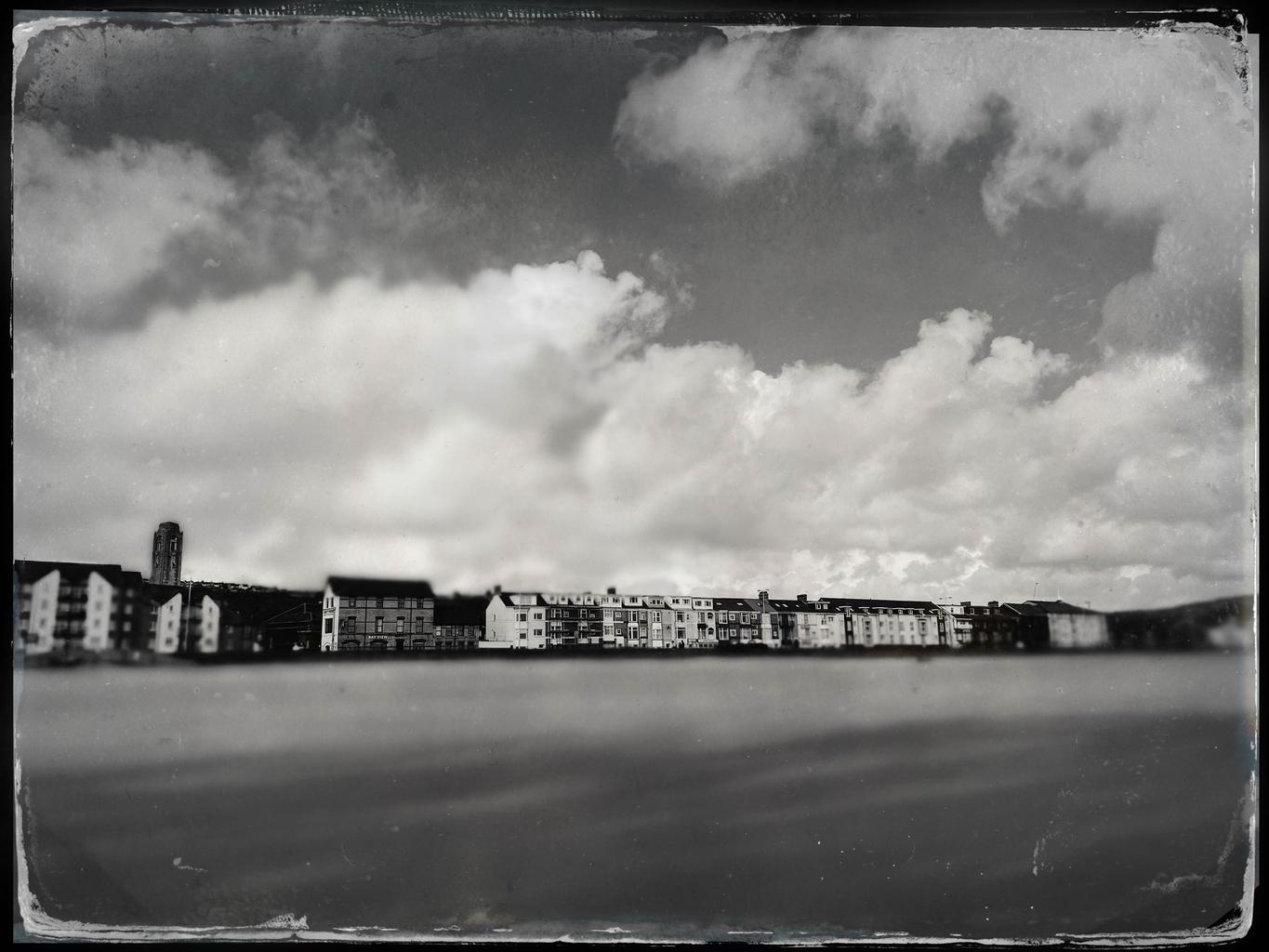 A black and white photo of houses along Oystermouth Road. Taken from Swansea beach, the houses and many of the clouds are in focus, but the beach is windblown and blurry.